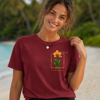 Une jeune femme souriante avec des taches de rousseur portant un t-shirt bordeaux de la marque VIROR sur une plage. Le t-shirt présente une illustration d'orchidée jaune et orange style aquarelle dans un cadre doré sur la poitrine gauche.